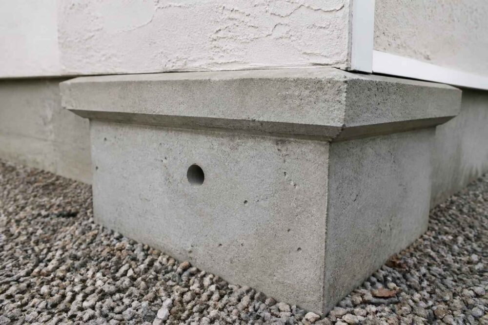 Betonowa podstawa z małym okrągłym otworem na żwirowym podłożu przy elewacji budynku