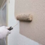 Dłoń w rękawiczce maluje zewnętrzną ścianę budynku wałkiem malarskim